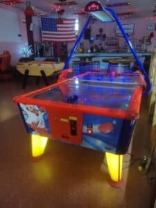 Table d'air hockey lumineuse rouge et bleue dans une salle de jeux. Drapeau américain, baby-foot, bar et guirlandes lumineuses. Prix 2€.