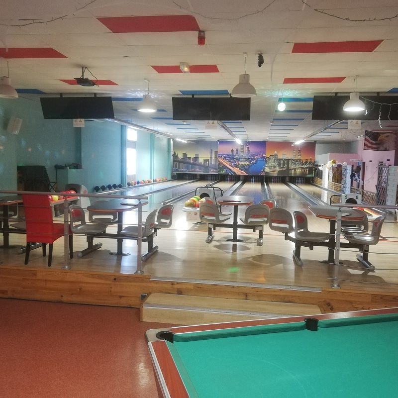 Bowling lumineux avec plusieurs pistes, un billard vert en avant-plan et des tables de repos. Murale urbaine lumineuse en fond.