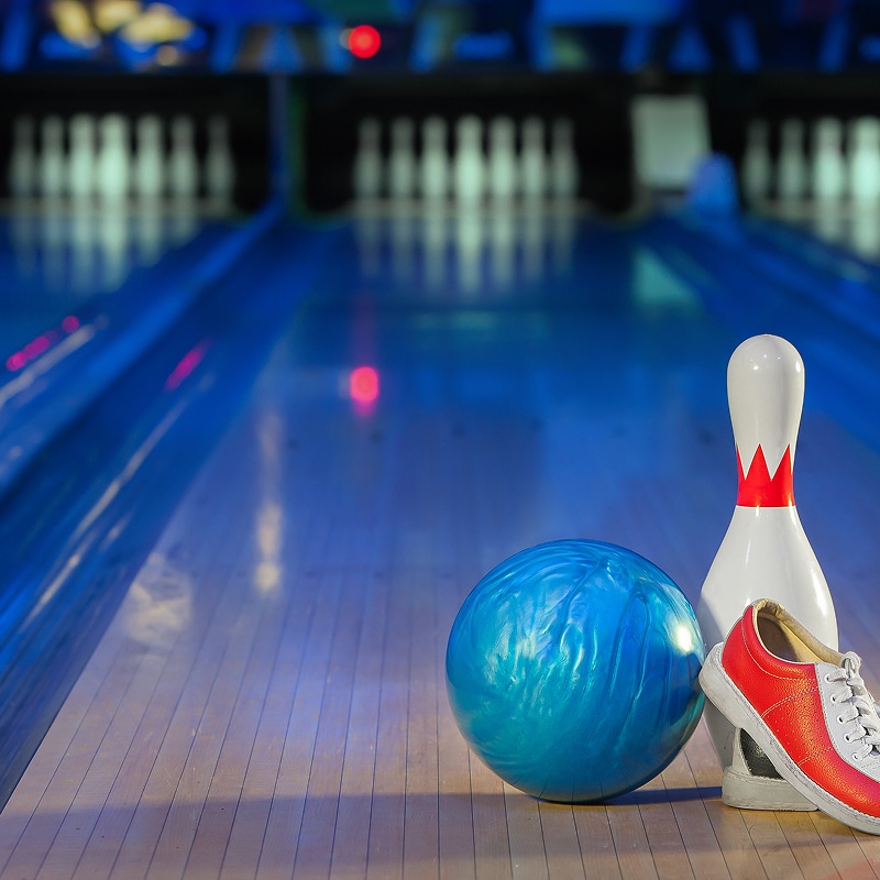 Bowling : Boule, quille et chaussure sur piste bleue Boule de bowling bleue, quille blanche et chaussure rouge posées sur une piste illuminée de bleu, pins au fond.