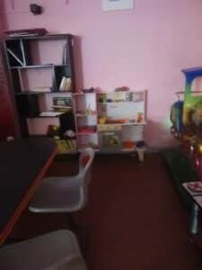 Salle de jeu enfantine aux murs roses. Une étagère de livres côtoie une cuisine jouet et un train coloré. Table et chaise visibles.