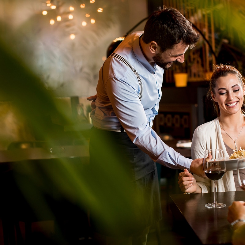 Service impeccable : Serveur souriant au restaurant Serveur souriant apportant un plat à une cliente joyeuse attablée avec un verre de vin rouge dans un restaurant chaleureux.