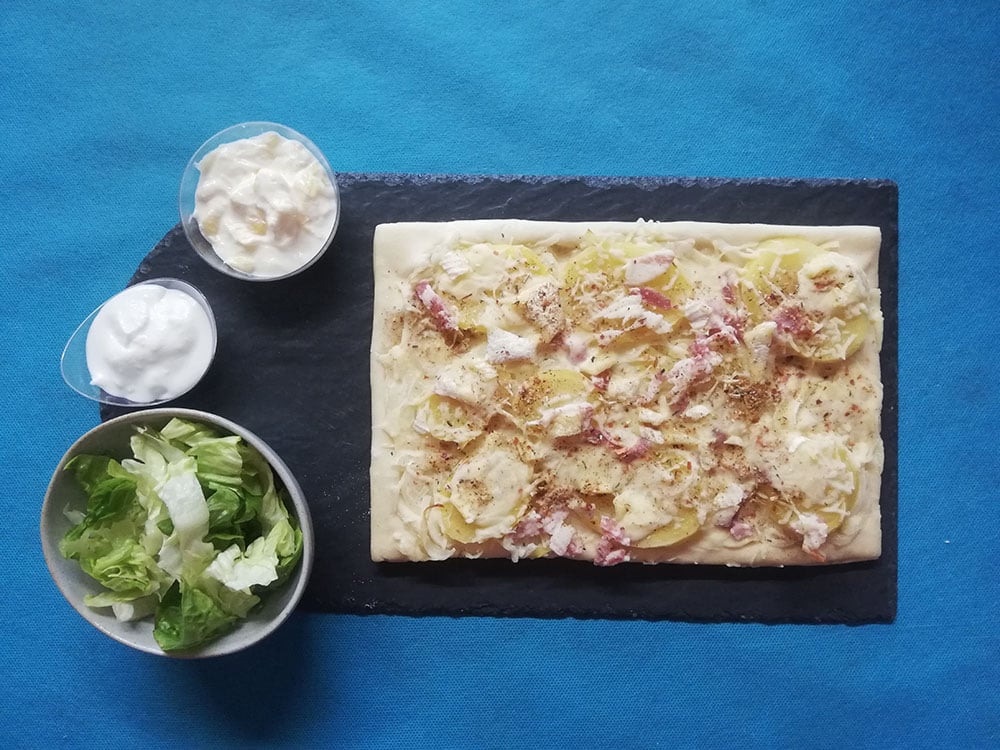 Tarte aux pommes de terre, lardons et oignons sur ardoise, servie avec une salade verte et deux sauces crémeuses.